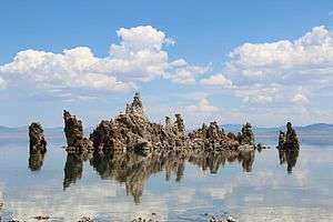 A rock formation rises from the surface of a calm lake. In the distance, the horizon is filled with hills.  The sky is blue with clouds The rocks appear grey in colour.