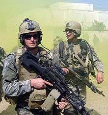 Michael Monsoor, a U.S. Navy SEAL, with a fellow SEAL team-mate, dressed in green camouflage uniform loaded with green combat uniforms. Both are carrying firearms and wearing sunglasses. There is a white-colored building and green smoke billowing in the background