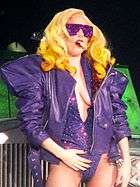 Lady Gaga in a big shouldered purple jacket and sunglasses, singing