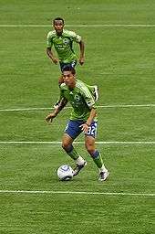 Two men in blue and green soccer uniforms. One has the ball at his feet and is looking upfield.