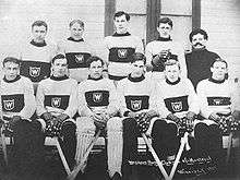 Black and white image of eleven men posing for a photo, with six of them sitting on a bench, and the other five standing behind them. All but one man are wearing the same wool sweater, white with a dark band around the torso; within the band is a small white "W" surrounded by a white square. The men on the bench are clearly holding hockey sticks, and the man third from the left is wearing padding on his legs. The man not wearing a sweater, standing on the far right, is simply wearing a dark shirt.