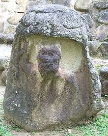 A standing stone with a large carved niche apparently representing the mouth of a monstrous creature, with its eyes carved onto the upper portion of the monument. The niche is occupied by a badly eroded figure depicted from the waist up. The figure has its right arm raised and with traces of a helmet on the figure's head.