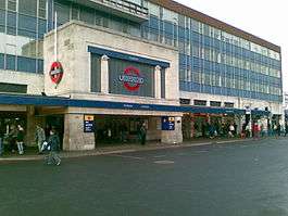 Station entrance in the form of a white stone-clad box sitting on two substantial and wide stone blocks. The front facade of the box contains a large London Underground logo (red ring with blue horizontal bar across the centre containing the word "UNDERGROUND") in the centre. Set back behind the entrance and to both sides a four-storey office block with blue cladding rises up.