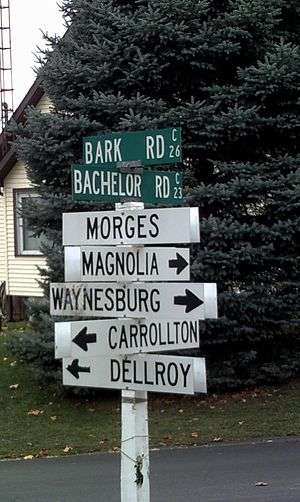 Signpost in front of a pine tree with seven signs on it reading from top to bottom Bark Road, Bachelor Road, Morges and four signs with arrows pointing different directions to Magnolia, Waynesburg, Carrollton, and Dellroy