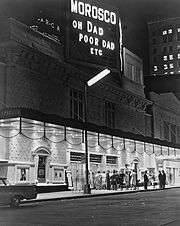 Night. Plain-looking theatre front from across the street, at an angle. Under the marquee, which is as wide as the theatre, a dozen or so people standing in front of the entrance are in bright light. Atop the building, a large sign announces the theatre's name and the play's name (abbreviated as Oh Dad Poor Dad, etc.). Everything else above the marquee, including nearby and distant buildings, is in relative darkness, but the street lights and illuminated signs and windows make it  visible.
