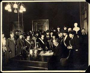 A man sits at a desk sigining a document while a large group of women watches