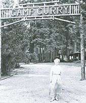 Woman in a dress in front of a sign above a road made of wood lettering. The sign reads "Camp Curry" and trees are in the background.
