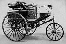 A black-and-white photograph shows a small open-air vehicle with three wheels, each spoked like bicycle tires. Visible are a small seat, a crank for steering, and a hand throttle for acceleration.