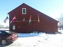 A red barn displaying the flags of Austria, Hungary, New Jersey, and the United States, with snow on the ground and a car in front.