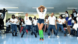 A woman that wears a white blouse, blue shorts, green socks, and red sneakers is standing. Behind her many people, similarly dressed, are dancing in the room.