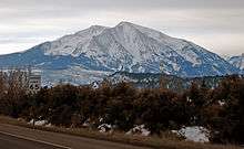 A snow-covered peak under a low overcast sky, seen from a road with bare brown vegetation in the foreground. To the left is a sign giving the speed limit as 65 miles per hour