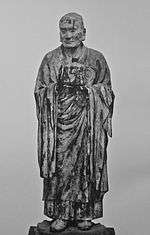 Muchaku (Asanga). Frontal view of a lifelike statue keeping his hands in front of his body with one palm turned up and the other facing sidewards as if keeping something. Black and white photograph.