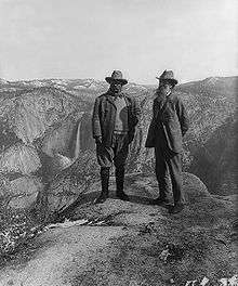 Two men stand at a precipice overlooking a valley that has a waterfall in the background.