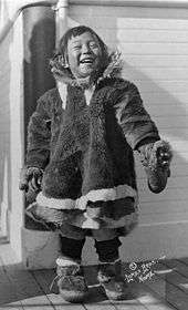 A small child standing on what appears to be a ship's deck. She is wearing a fur smock and fur boots, and has an open, happy expression.