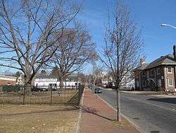 A color photo. Down the middle runs a tree-lined sidewalk, with a fenced grassy area to the left and a street to the left, lined with houses.