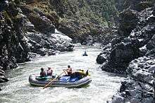 Two rafts, one with three people and one with one person, negotiate fast-moving water in a narrow, rocky river canyon.