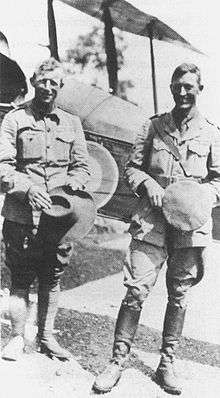 Full-length portrait of two men in military uniforms, standing in front of a biplane