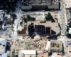 An overhead view shows the Alfred P. Murrah building, half of it destroyed from the bomb's blast. Near the building are various rescue vehicles and cranes. Some damage is visible to nearby buildings.