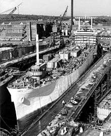 A large ship sits in drydock at the yard, with cranes in the background