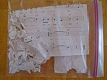 A ziplock plastic bag on a wooden surface containing shreds of paper with musical notes and a staff on them