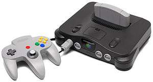 The Nintendo 64 controller is light gray controller with three handles for the player's two hands. It has red, green, blue, and yellow buttons, an analog stick, and a directional pad. The controller is plugged into the charcoal gray Nintendo 64 with a light gray cartridge inserted. The sleek console is convex on its top, and has two power switches and four controller ports.