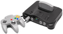 On the left, a three-pronged, handheld controller with a central analog stick and multiple buttons. On the right, a black electronics unit that accepts a light gray cartridge on the top and controllers (via ports) on its front.