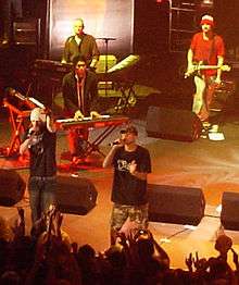 All-male group on stage, containing two MCs, two keyboardists, and a guitarist. Some of the crowd with hands in the air present in the foreground