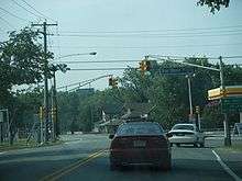 A two lane road at a traffic light in a built up area. Two traffic light poles are located on each side of the road, with a green sign on the left pole arm reading left arrow Burleigh Avenue and a green sign on the right pole arm reading Bay Shore Road County Route 603 right arrow