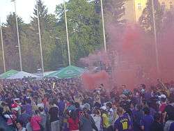 Hundred of fans, wearing purple, celebrating with purple smoke from flares in the background.