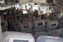 Interior of the aircraft, after the crash, showing oxygen masks hanging from the ceiling, ready for use. The seat backs have video displays installed, and most seats are in position, some reclined, some upright. The overhead compartments are open.