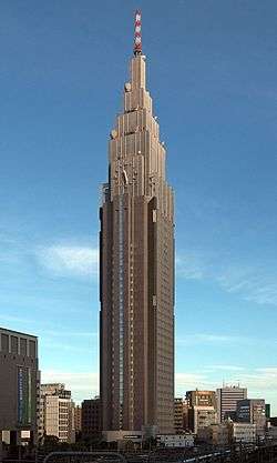 Ground-level view of a brown, rectangular high-rise; as it rises, it terraces to a point and a white and an orange antenna rises from the top. A clock is located on one side of the building.