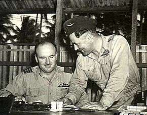 Two men in military uniforms conversing at a desk in a wooden hut