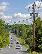 A road, with wooden telephone poles and lines on either side, descending from the camera into a wooded area and then ascending in the background. Cars are traveling it in both directions.