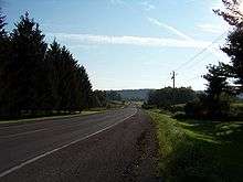 A two-lane highway heads through an area containing open fields and areas of trees. A large, tree-covered ridge is in the distant background.