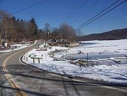 A road winding by the shore of a frozen lake and some docks in wintertime. There are telephone lines over the road, light snow on the ground, and a wooded hill on the far side of the lake at the right of the image