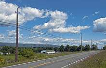 A two lane road cutting across a gentle slope seen from just above under a blue sky with large clouds. Across the road are telephone poles with lines and just downhill is a small farm building, white with a green roof and horses and cows in the vicinity. In the distance a wooded ridge rises.