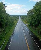 A wet paved road, seen from above. It continues straight ahead into the center of the image, leaving a narrow notch in the foreground to enter a wide, flat landscape in the rear.