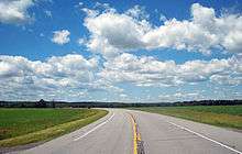 A paved two-lane road curving to the left into the distance through a large grassy area below a blue sky filled with cumulus clouds