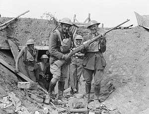 A black and white photograph of a group of men in military uniform outside a dugout in a trench, with one man helping another display a gun for the camera