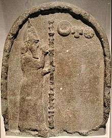 Stone stele with a carving depicting a man with a beard, carrying a tall staff and wearing a robe and conical hat, gesturing to three symbols representing the moon, sun and Venus.