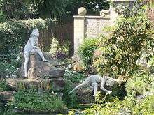 Colour photograph, zoom shot through trees of two nude female statues on a rockery, one sitting, the other in a strange pose