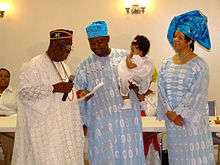 An Igbo couple standing with the dad holding a what looks to be a girl child in a naming ceremony, a man known as di okpara or the head of the family, standing left, checks the names with the couple looking one. The wear blue lace outfits and the di okpara wears white. The baby wears white, the woman wears a blue head tie and the father a blue cap, the di okpara wears a red cap