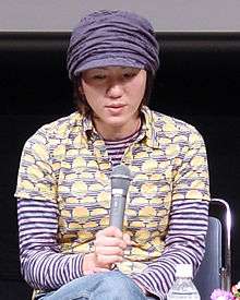 A Japanese woman wearing a newsboy cap and patterned shirt, holding a microphone.