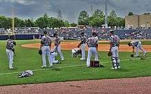 Baseball players wearing white pants, olive and tan camouflage jerseys, and black hats, on a green baseball field stretching before a game.