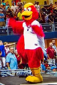 A person wearing a red anthropomorphized rooster costume dressed in a white baseball jersey with a blue "V" on the right chest dances on a baseball dugout