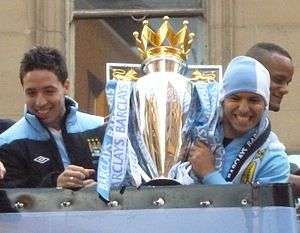 Two young men dressed in sportswear hold a large trophy aloft. The man on the right has a blue and white scarf wrapped around his head.