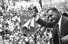 A black and white image of a man at left waving to a crowd behind him in the rest of the image that is waving Egyptian flags and holding a picture of him