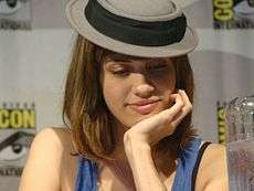 A brown-haired woman wearing a gray hat and blue tank top shirt leans her face into her left hand.
