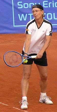 A blond-haired women with a white shirt, black shorts, and white tennis shoes on about ready to serve the tennis ball in hand