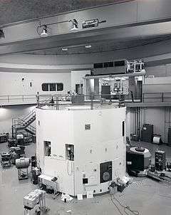 The black-and-white photograph is of a large room that contains a lot of electronic equipment. The lower half of the image contains a cylindrical white container that is a nuclear reactor. There is a walkway at the top of the reactor, which leads back to a control room where two men are sitting.